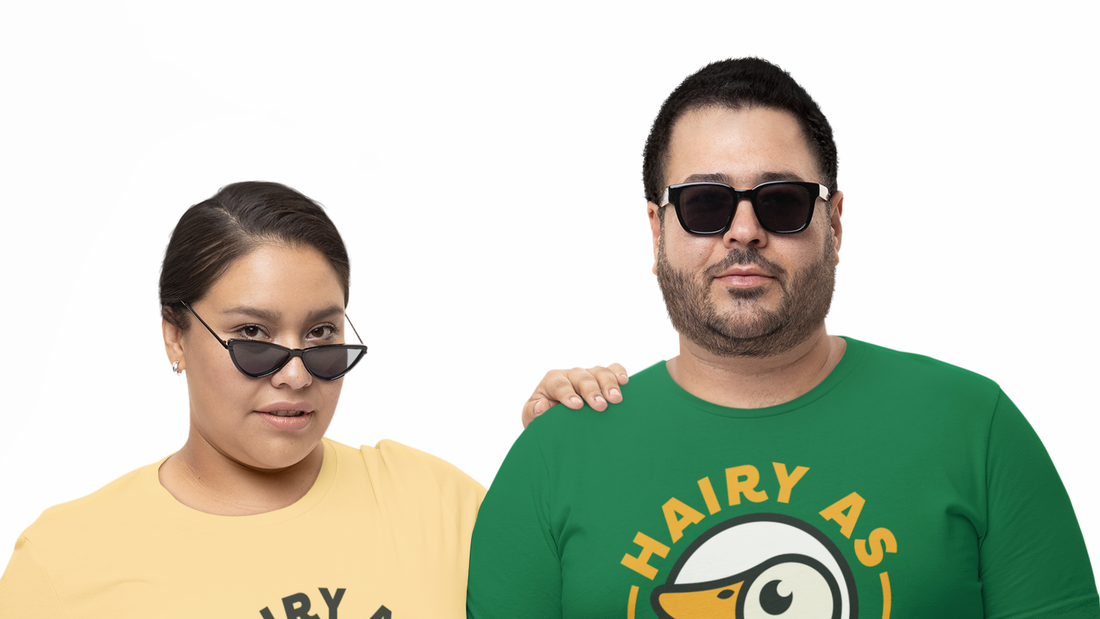 Man on the right has a short beard wearing, is wearing sunglasses, and has a green Hairy As t-shirt on. Woman on the left has her sunglasses on her nose and is wearing a mustard Hairy As t-shirt.