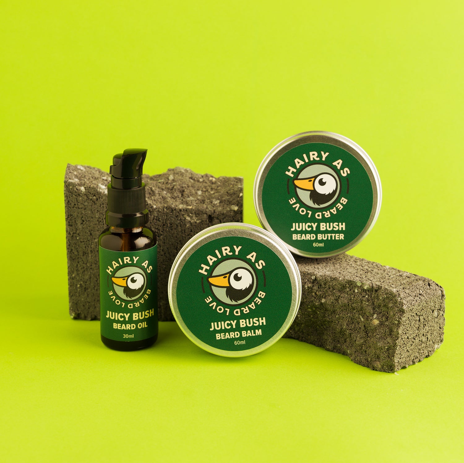 Hairy As's Australian-made Juicy Bush Beard Collection. An amber 30ml bottle of Beard Oil, and 60ml aluminium tins of Beard Butter and Beard Balm. Labels are green with jasmine font. They are displayed arranged on some cement bricks on a green background.