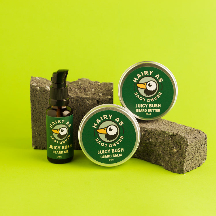 Hairy As's Australian-made Juicy Bush Beard Collection. An amber 30ml bottle of Beard Oil, and 60ml aluminium tins of Beard Butter and Beard Balm. Labels are green with jasmine font. They are displayed arranged on some cement bricks on a green background.