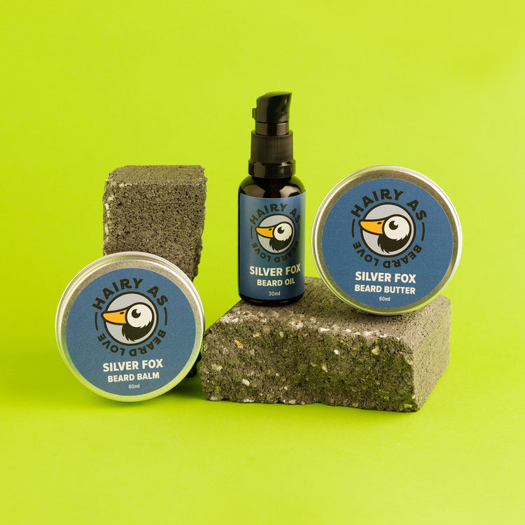 Hairy As's Australian-made Silver Beard Collection. An amber 30ml bottle of Beard Oil, and 60ml aluminium tins of Beard Butter and Beard Balm. Labels are blue with silver font. They are displayed arranged on some cement bricks on a green background.