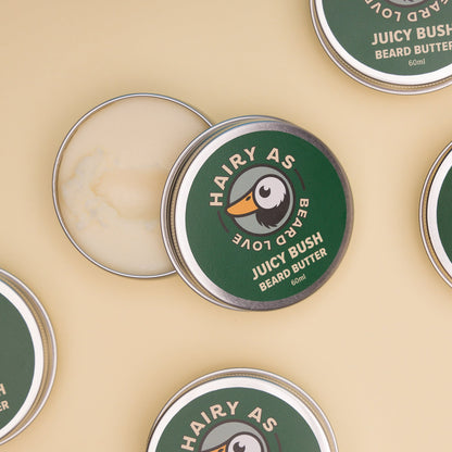 Hairy As Beard Love Beard Butter blend Juicy Bush. A 60ml grey aluminium screw top tin that is open so you can see inside. Four additional tins visible on the edge of frame. Label is green with cream coloured font. Features the Hairy As logo of a bearded duck.