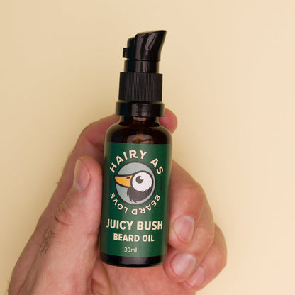 Hairy As Beard Love Beard Oil blend Juicy Bush. A 30ml amber glass bottle with a black serum pump cap. Label is green with cream coloured font. Being held in a left hand. Features the Hairy As logo of a bearded duck. Jasmine background.